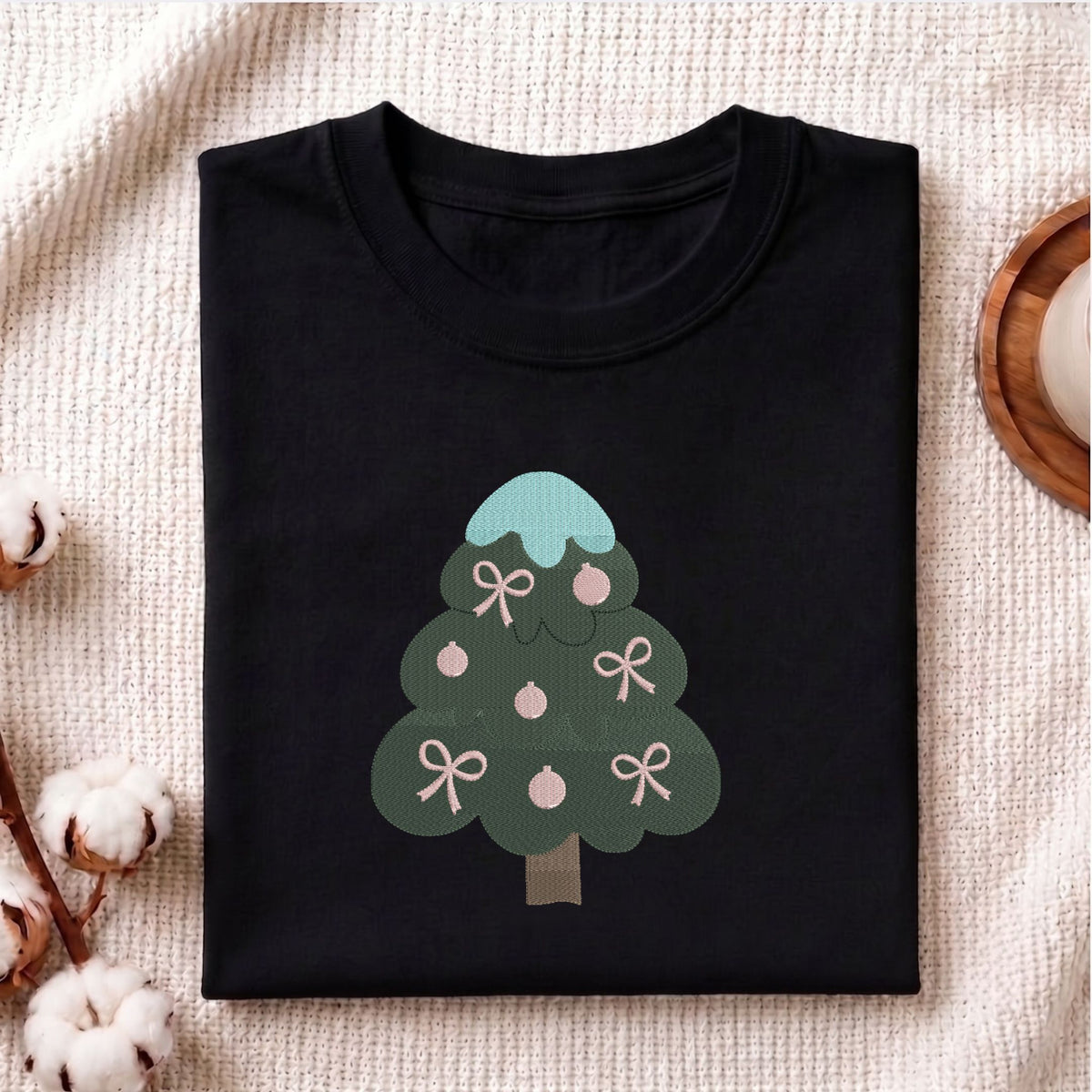Folded black cotton t-shirt mockup featuring a Christmas tree machine embroidery design, styled with cotton stems and a wooden cup.