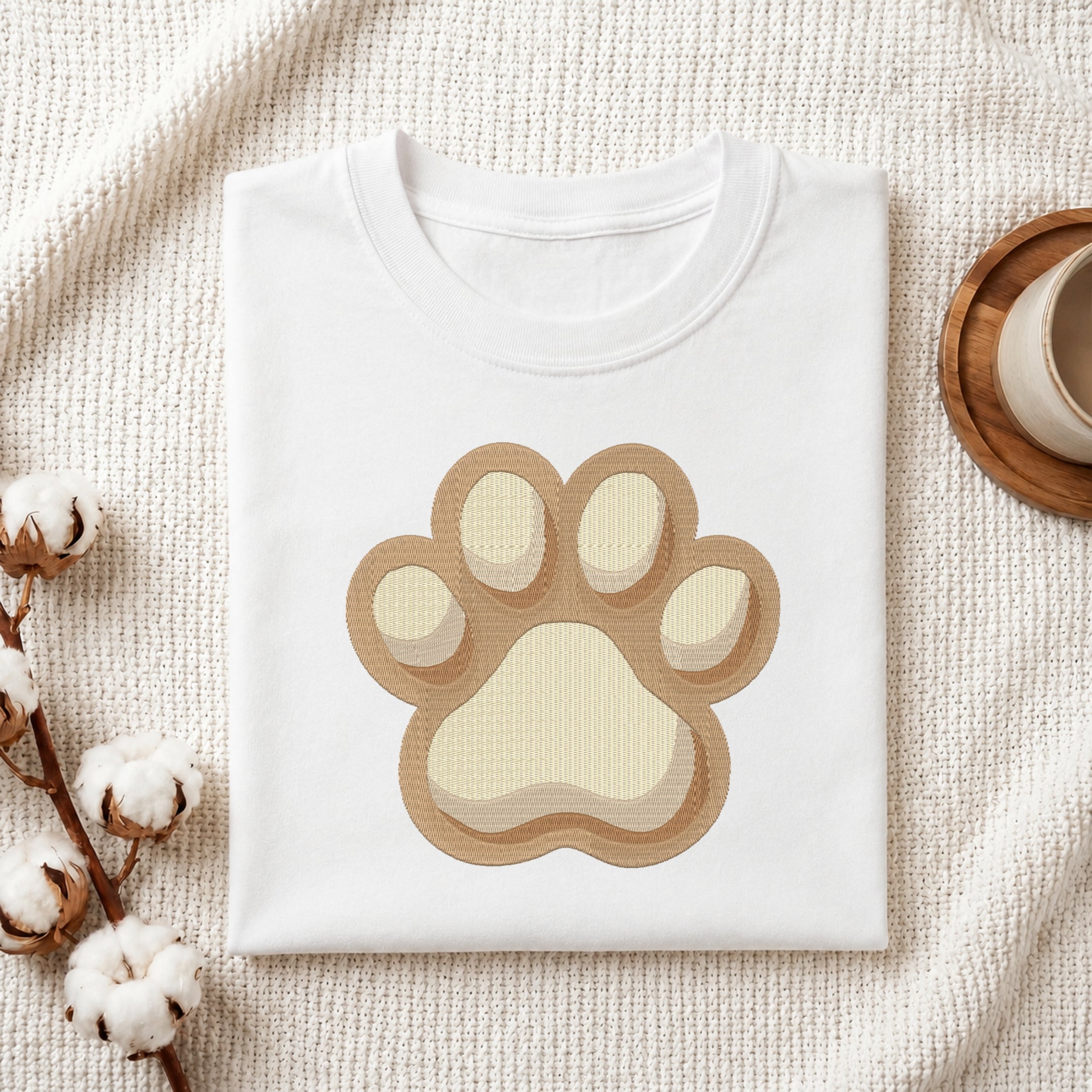 A folded white T-shirt with a large layered paw print graphic in tan and cream, styled on a textured cream fabric with cotton stems and a small cup at the side.