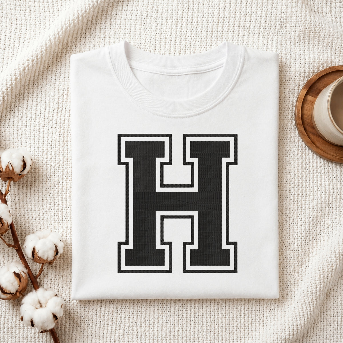 A folded white T-shirt with a large black block letter 'H' outlined in white on the front, styled on a textured cream fabric with cotton stems and a small cup at the side.