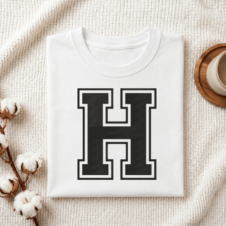 A folded white T-shirt with a large black block letter 'H' outlined in white on the front, styled on a textured cream fabric with cotton stems and a small cup at the side.