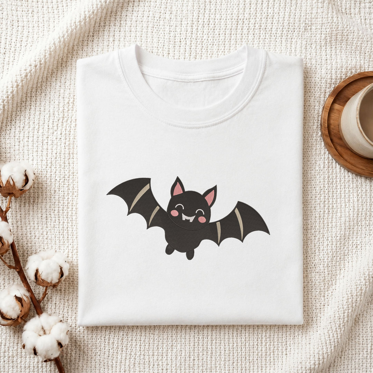 Folded white cotton t-shirt mockup featuring a cute Halloween bat machine embroidery design, styled with cotton stems and a wooden cup.