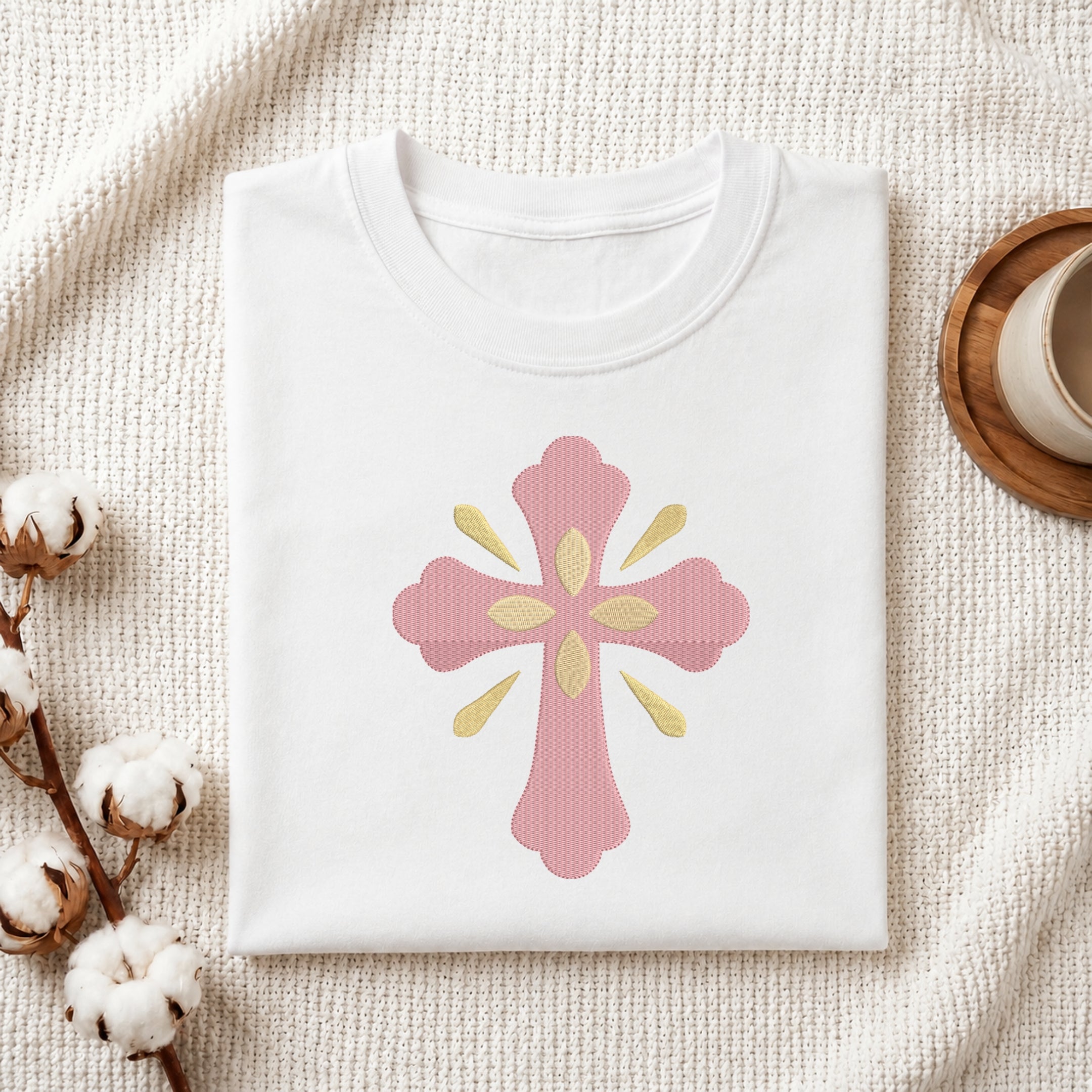 Folded white cotton t-shirt featuring a pink and gold ornamental cross embroidery design centered on the chest.