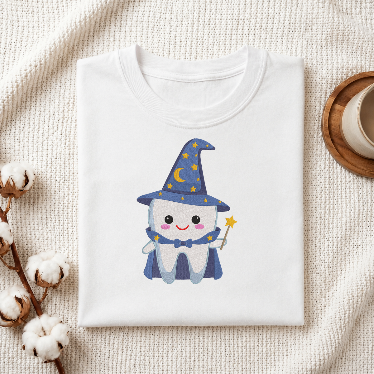 Folded white cotton t-shirt mockup featuring a cute wizard tooth machine embroidery design, styled with cotton stems and a wooden cup   
