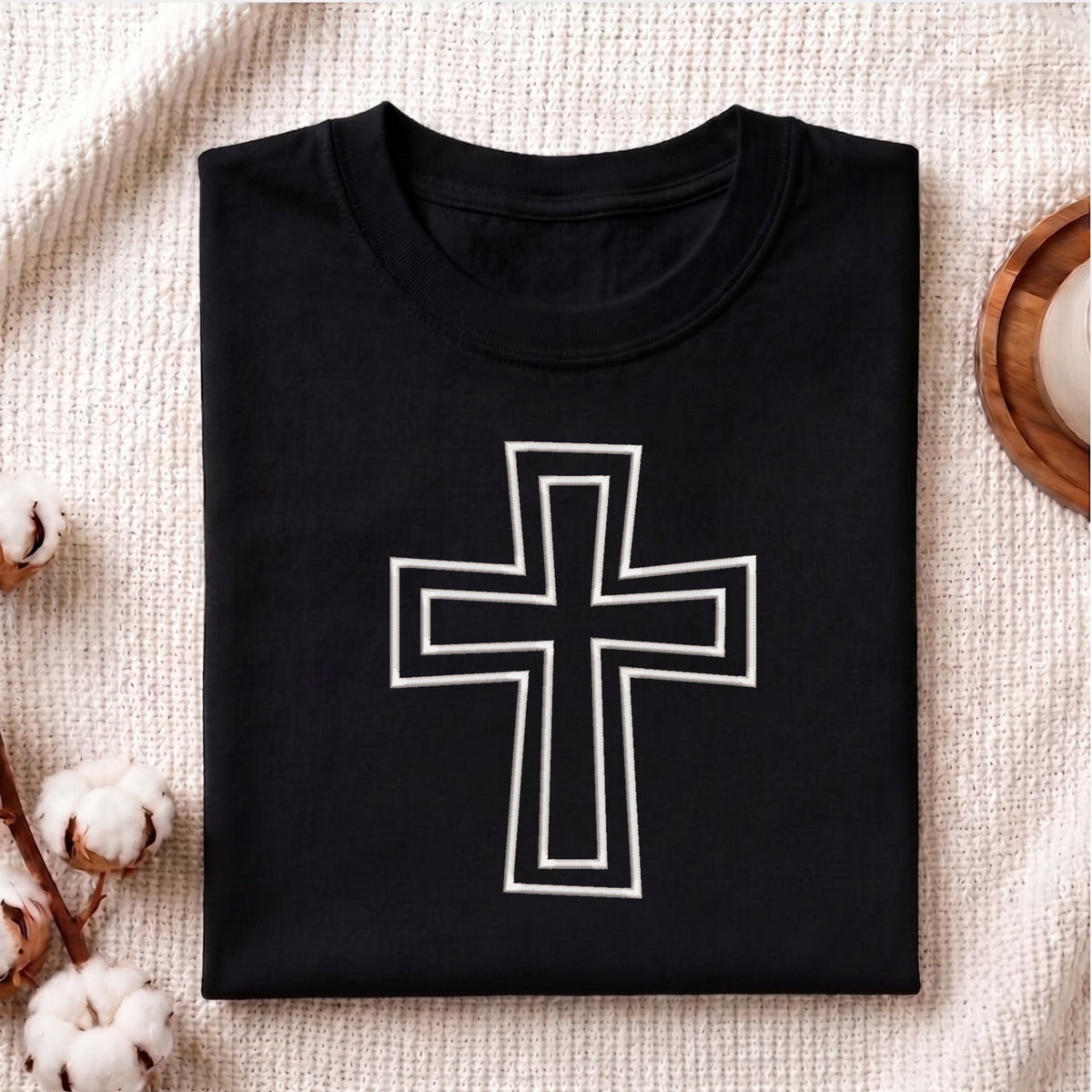 A neatly folded black crewneck t-shirt featuring the white outlined Christian cross embroidery design centered on the front, styled on a textured knit blanket next to cotton stems.