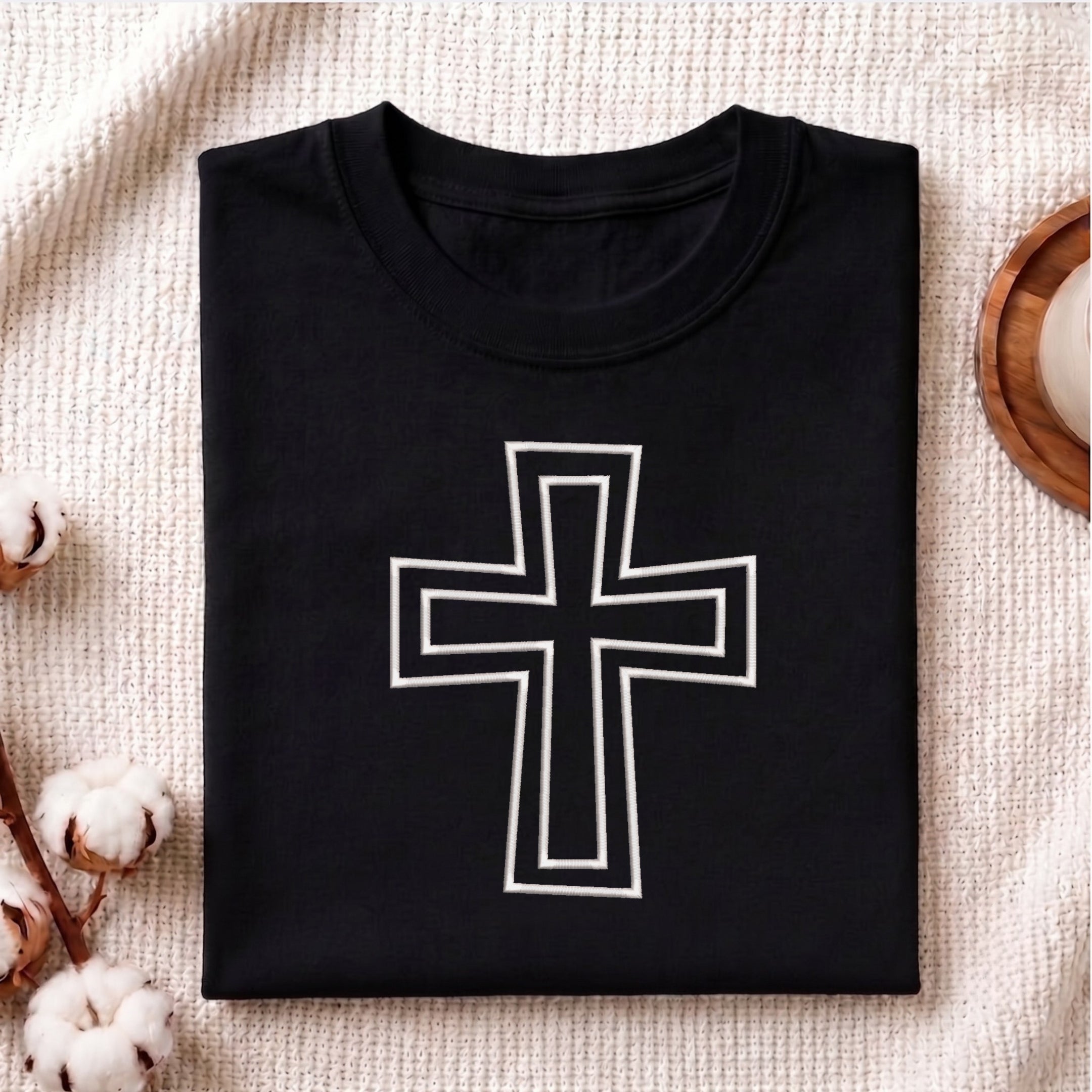 A neatly folded black crewneck t-shirt featuring the white outlined Christian cross embroidery design centered on the front, styled on a textured knit blanket next to cotton stems.