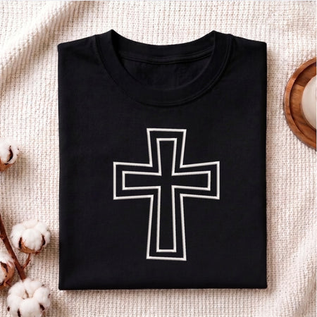 A neatly folded black crewneck t-shirt featuring the white outlined Christian cross embroidery design centered on the front, styled on a textured knit blanket next to cotton stems.