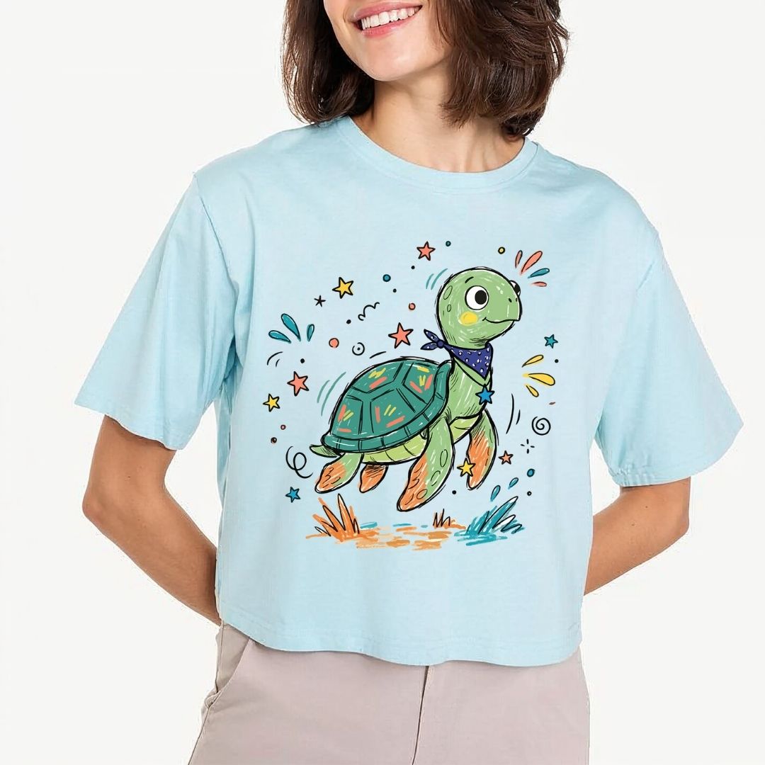 A woman wearing a light blue cropped t-shirt with a colorful cartoon sea turtle graphic on the front, paired with beige pants, against a plain white background.
