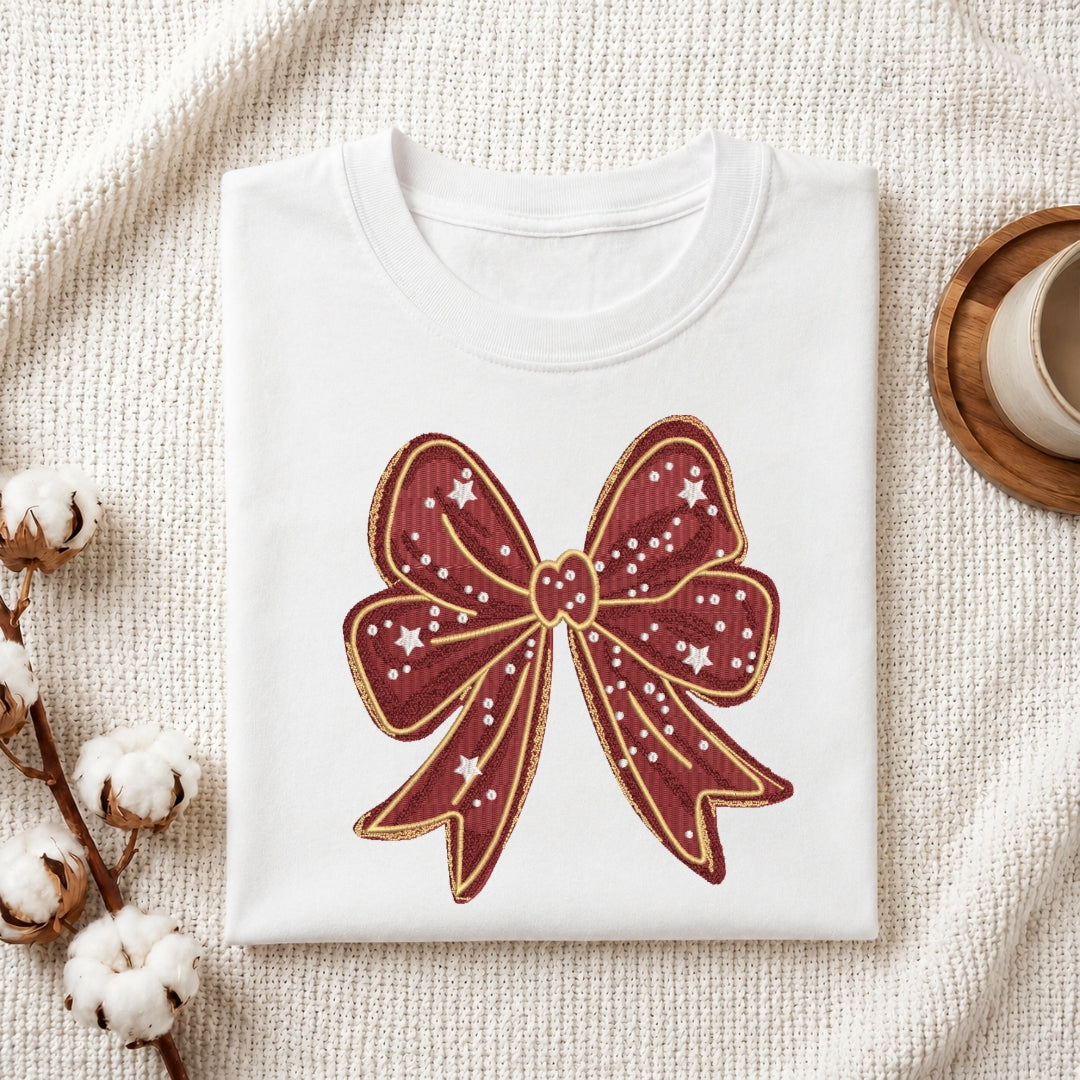 A folded white crewneck t-shirt featuring a large embroidered burgundy bow with gold outlines and white star details, styled on a textured cream knit blanket with cotton stems and a wooden cup in the background.