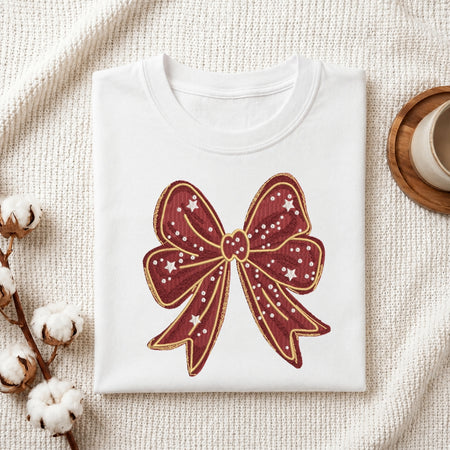 A folded white crewneck t-shirt featuring a large embroidered burgundy bow with gold outlines and white star details, styled on a textured cream knit blanket with cotton stems and a wooden cup in the background.