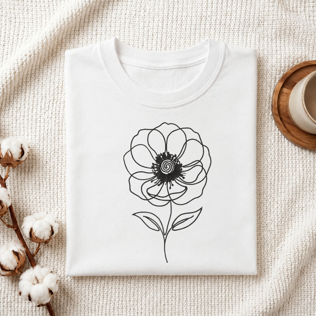 A folded white crewneck t-shirt with a black line art flower graphic on the front, styled on a textured cream knit blanket with cotton bolls and a wooden cup in the background.