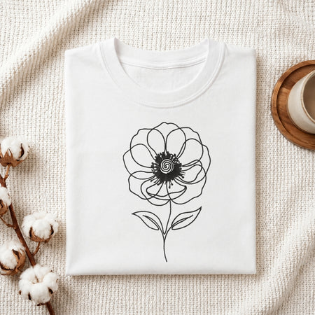 A folded white crewneck t-shirt with a black line art flower graphic on the front, styled on a textured cream knit blanket with cotton bolls and a wooden cup in the background.