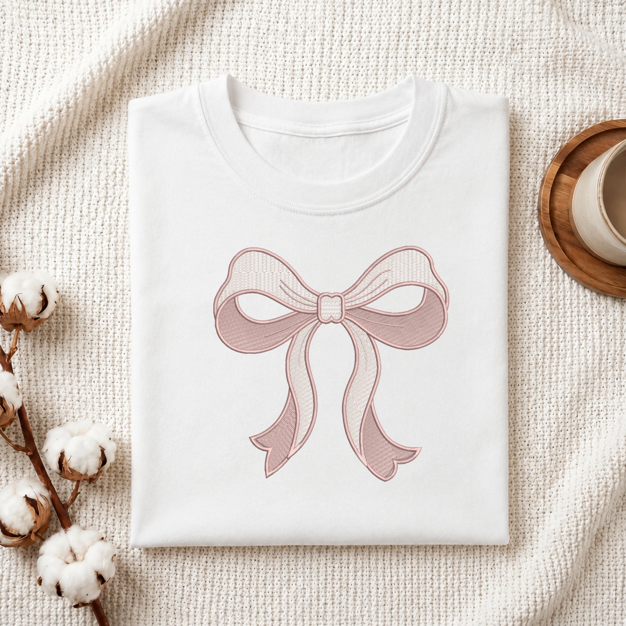 A folded white T-shirt with a pink bow graphic printed on the front, styled on a textured cream fabric with cotton stems and a wooden cup in the background.