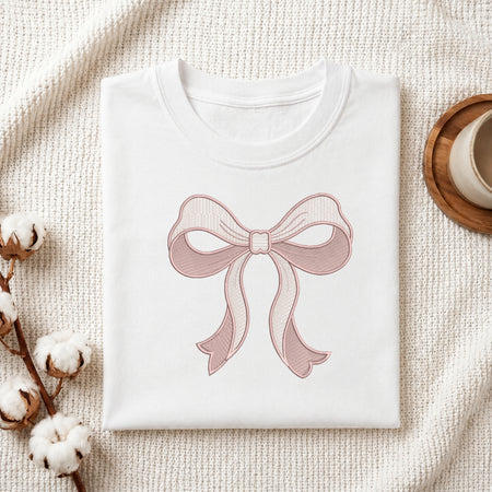 A folded white T-shirt with a pink bow graphic printed on the front, styled on a textured cream fabric with cotton stems and a wooden cup in the background.