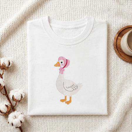 Folded white cotton t-shirt mockup featuring a cute goose wearing a pink bonnet machine embroidery design, styled with cotton stems and a wooden cup.