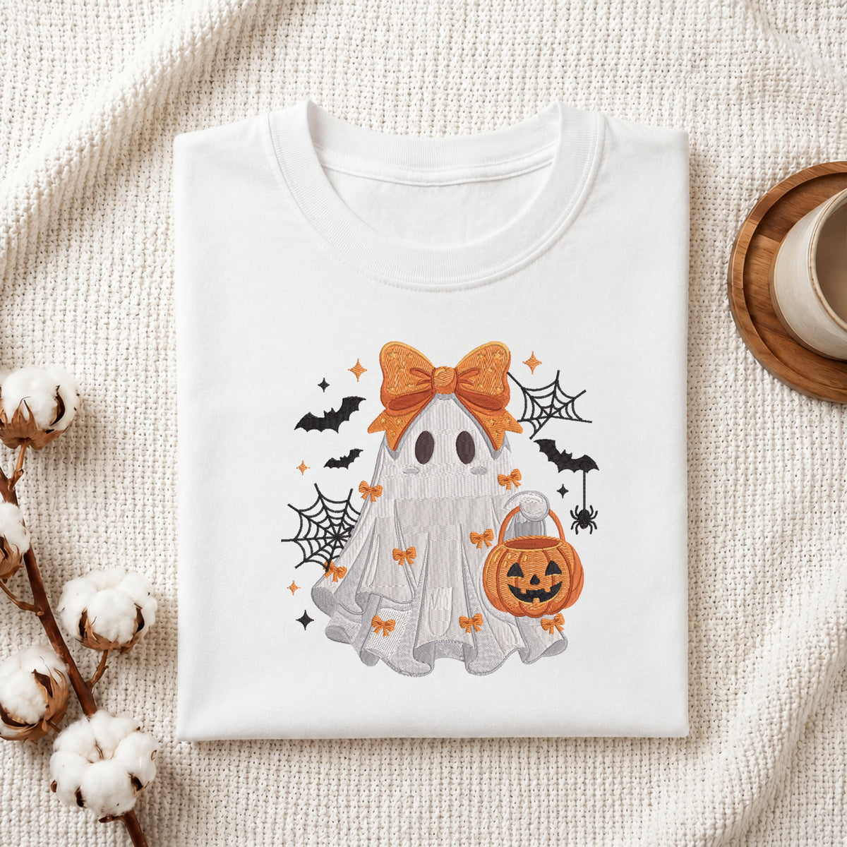 A neatly folded white t-shirt featuring the embroidered ghost with bow and pumpkin bucket graphic, displayed on a textured white knit background with cotton stems and a wooden cup.