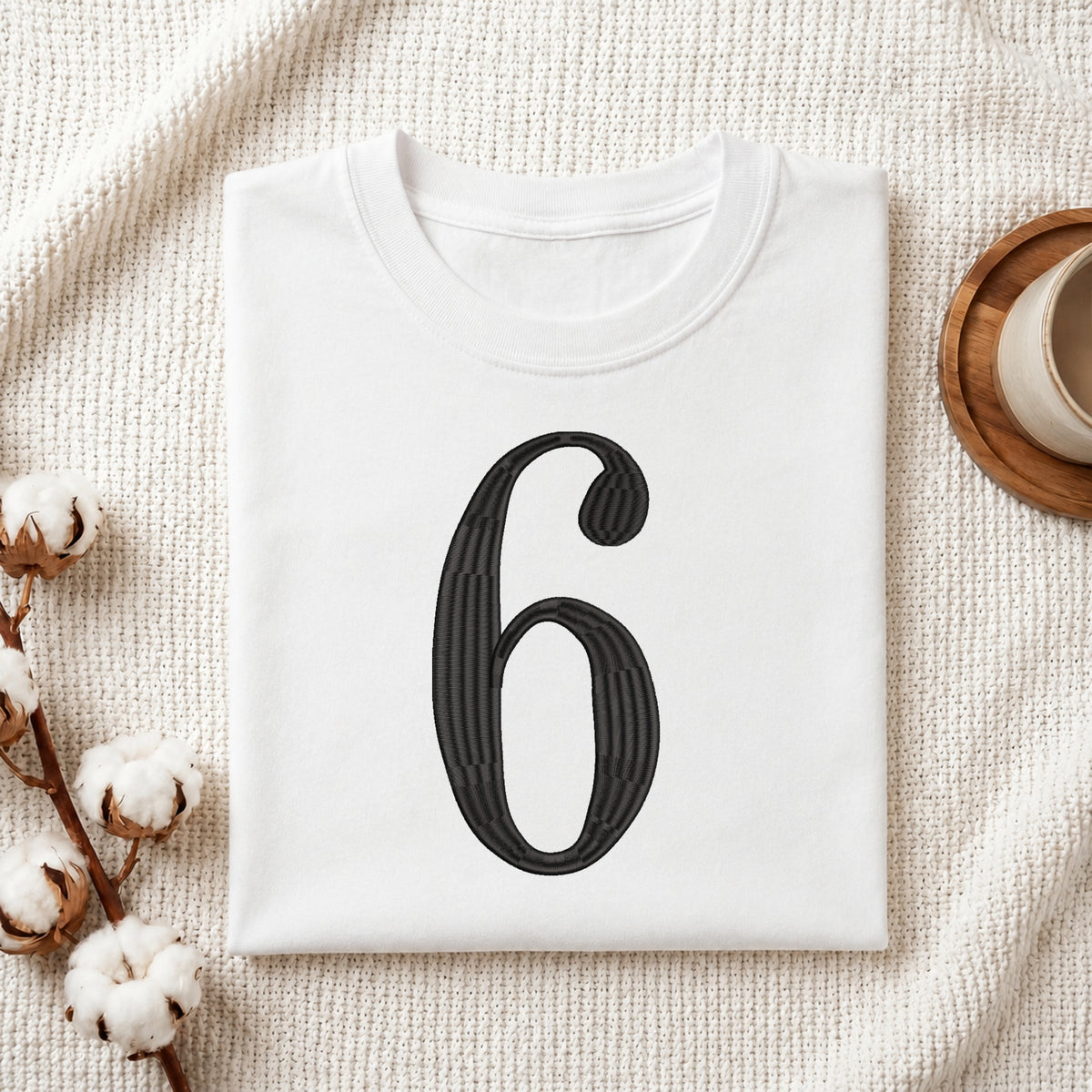 A folded white T-shirt with a large black number 6 printed on the front, styled on a textured cream fabric with cotton bolls and a small cup in the corners.