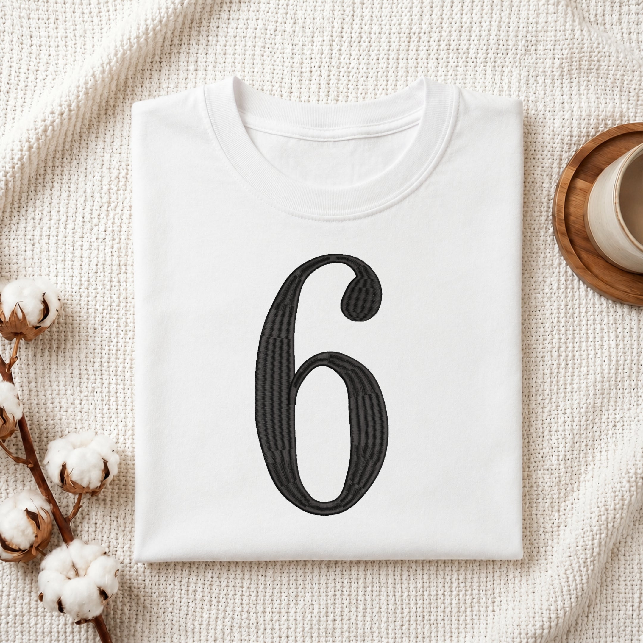 A folded white T-shirt with a large black number 6 printed on the front, styled on a textured cream fabric with cotton bolls and a small cup in the corners.