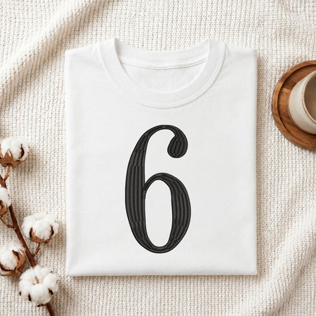 A folded white T-shirt with a large black number 6 printed on the front, styled on a textured cream fabric with cotton bolls and a small cup in the corners.