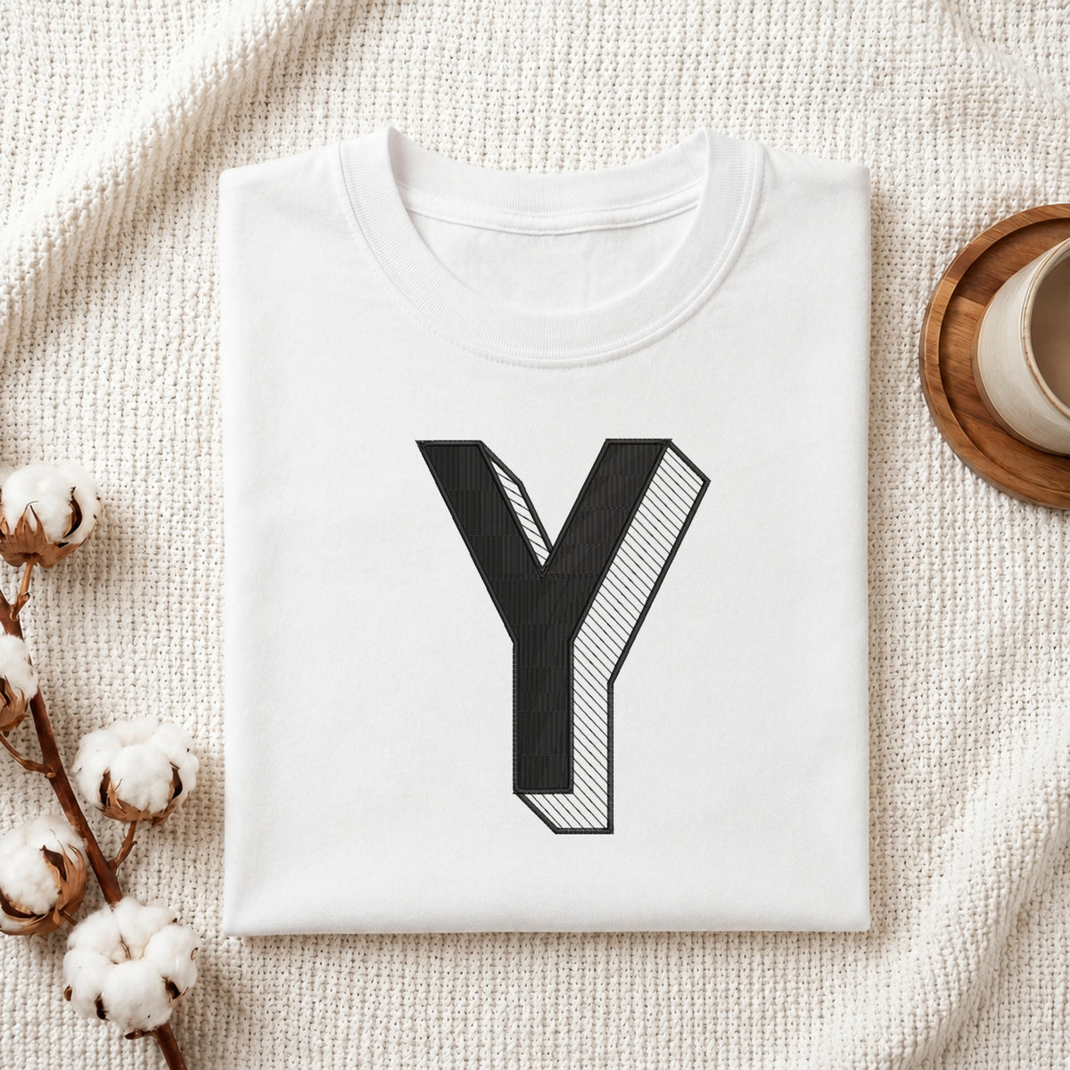 A folded white t-shirt with a large black graphic letter 'Y' printed on the front, styled on a textured cream knit blanket with cotton stems and a cup in the background.