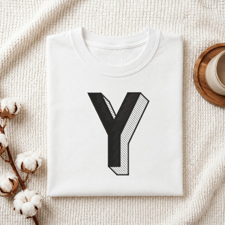 A folded white t-shirt with a large black graphic letter 'Y' printed on the front, styled on a textured cream knit blanket with cotton stems and a cup in the background.