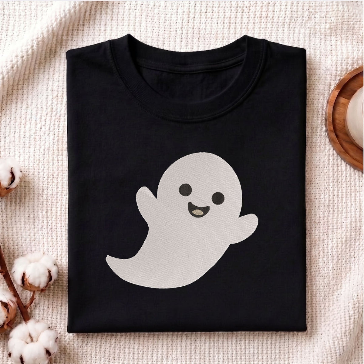Folded black cotton t-shirt mockup featuring a cute smiling ghost machine embroidery design, styled with cotton stems and a wooden cup.
