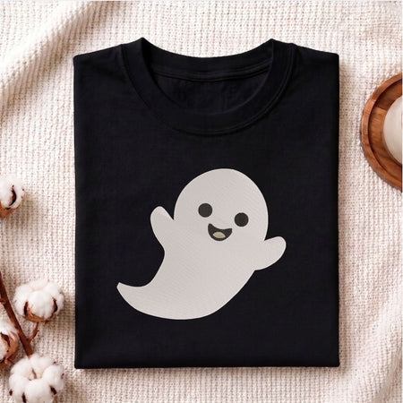 Folded black cotton t-shirt mockup featuring a cute smiling ghost machine embroidery design, styled with cotton stems and a wooden cup.
