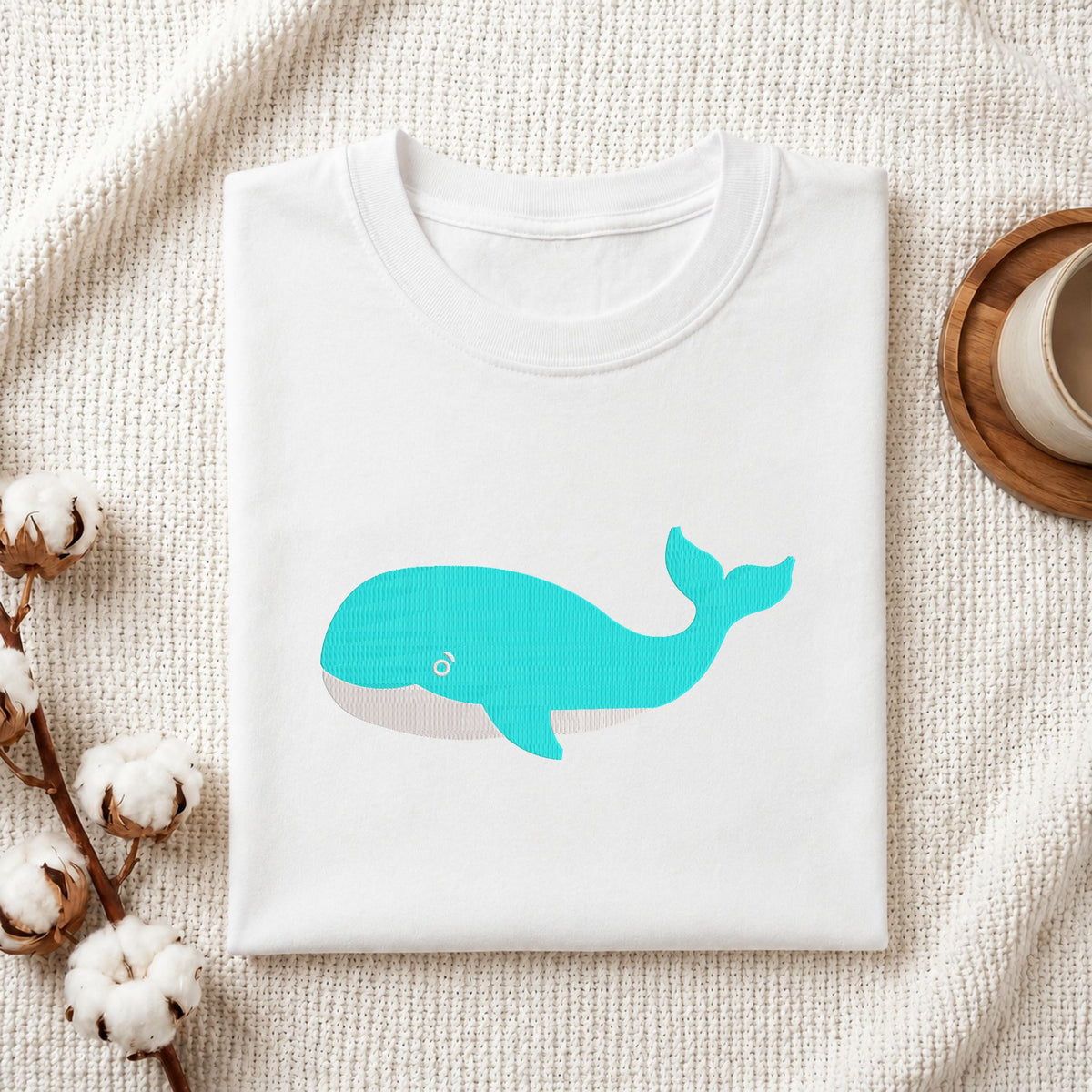Folded white cotton t-shirt mockup featuring a turquoise blue whale machine embroidery design, styled with cotton stems and a wooden cup.