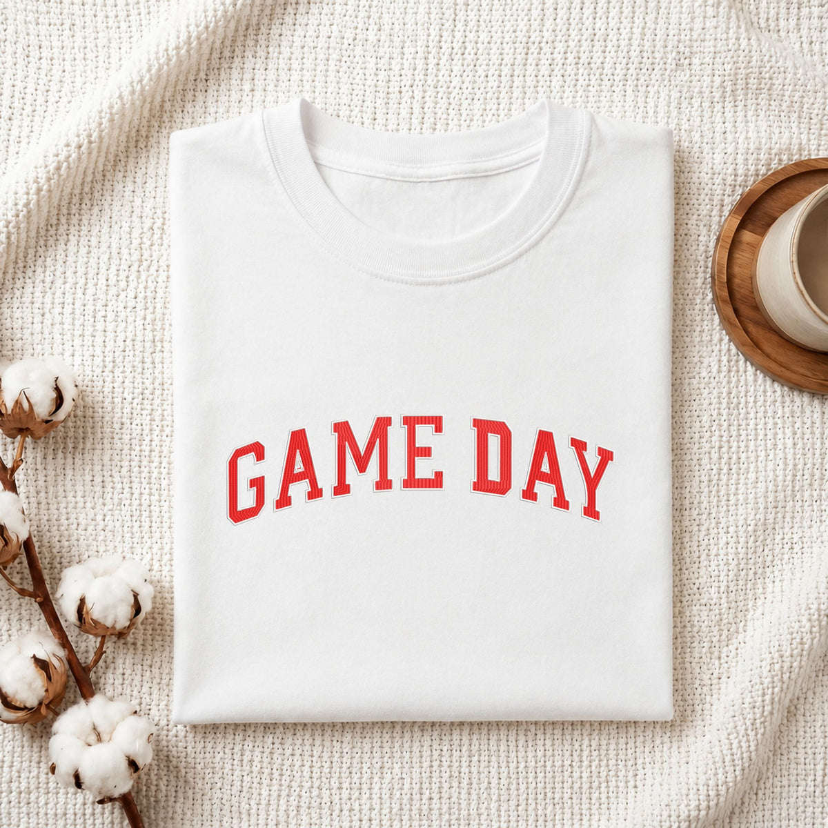 Folded white cotton t-shirt mockup featuring a red "GAME DAY" sports typography machine embroidery design, styled with cotton stems and a wooden cup.
