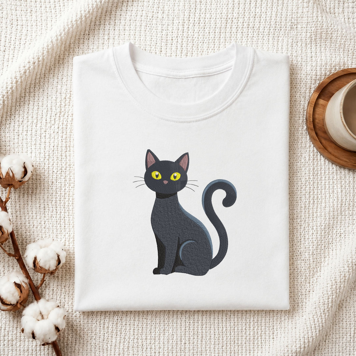 Folded white cotton t-shirt mockup featuring a cute sitting black cat machine embroidery design, styled with cotton stems and a wooden cup.