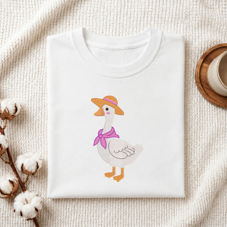 Folded white cotton t-shirt mockup featuring a cute duck wearing a straw hat and purple scarf machine embroidery design, styled with cotton stems and a wooden cup   