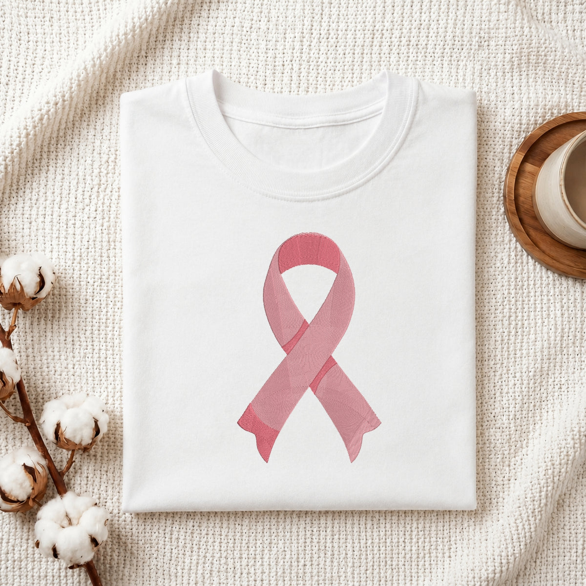 A folded white crewneck t-shirt featuring a pink watercolor-style awareness ribbon printed on the front, styled on a textured cream knit blanket with cotton bolls and a wooden cup in the background.