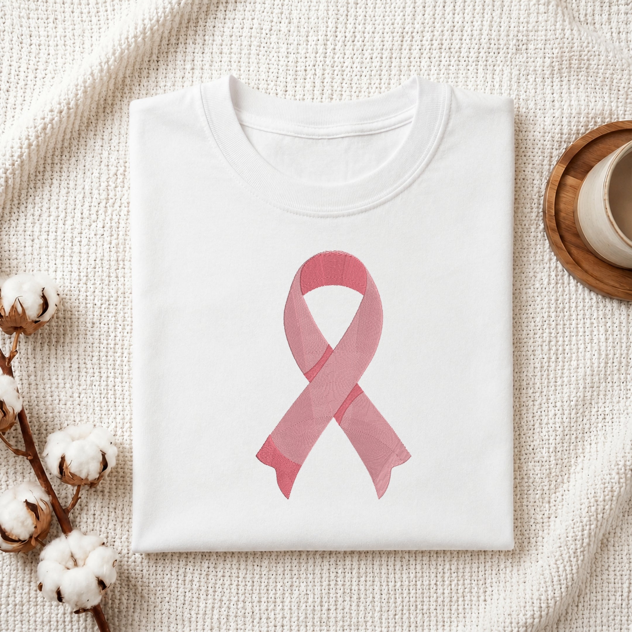 A folded white crewneck t-shirt featuring a pink watercolor-style awareness ribbon printed on the front, styled on a textured cream knit blanket with cotton bolls and a wooden cup in the background.