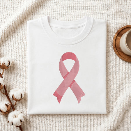 A folded white crewneck t-shirt featuring a pink watercolor-style awareness ribbon printed on the front, styled on a textured cream knit blanket with cotton bolls and a wooden cup in the background.