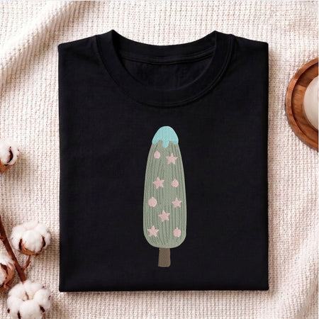 A folded black crewneck t-shirt featuring a graphic of a cactus-shaped popsicle with pink stars and a light blue top, displayed on a textured cream fabric with cotton bolls and a wooden coaster.