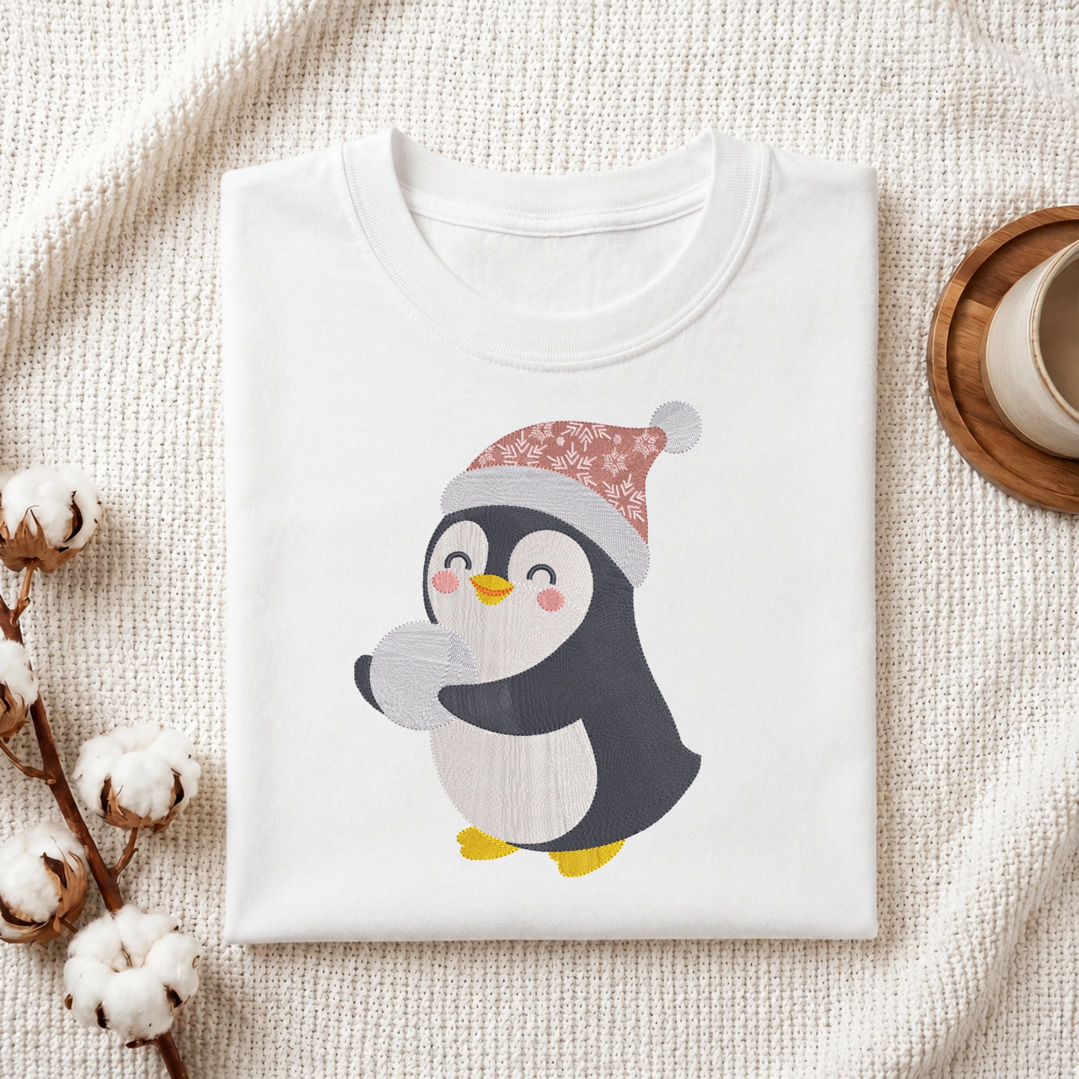 A folded white crewneck t-shirt featuring a cute cartoon penguin wearing a pink and gray patterned Santa hat, styled on a textured cream fabric with cotton bolls and a cup in the background.