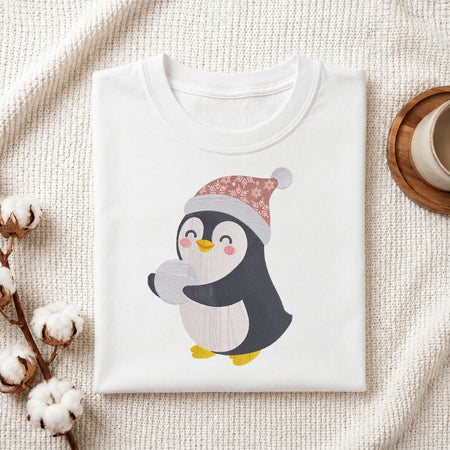 A folded white crewneck t-shirt featuring a cute cartoon penguin wearing a pink and gray patterned Santa hat, styled on a textured cream fabric with cotton bolls and a cup in the background.