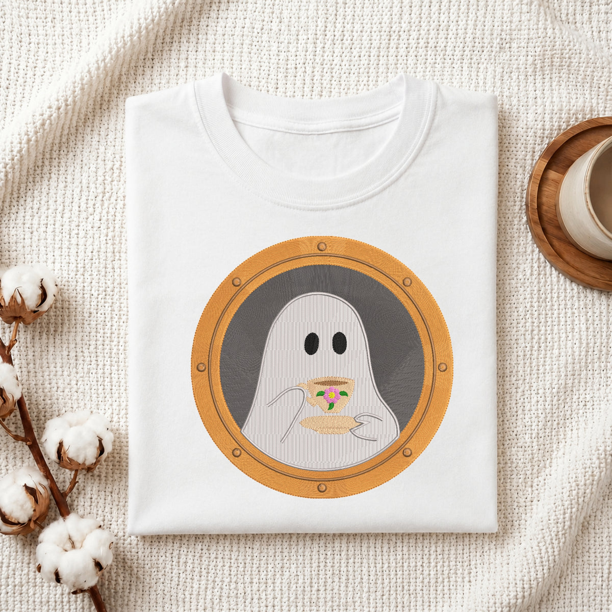 A folded white t-shirt with a graphic of a cute ghost eating a slice of pizza inside a porthole circle, styled on a textured cream blanket with cotton bolls and a coffee cup.