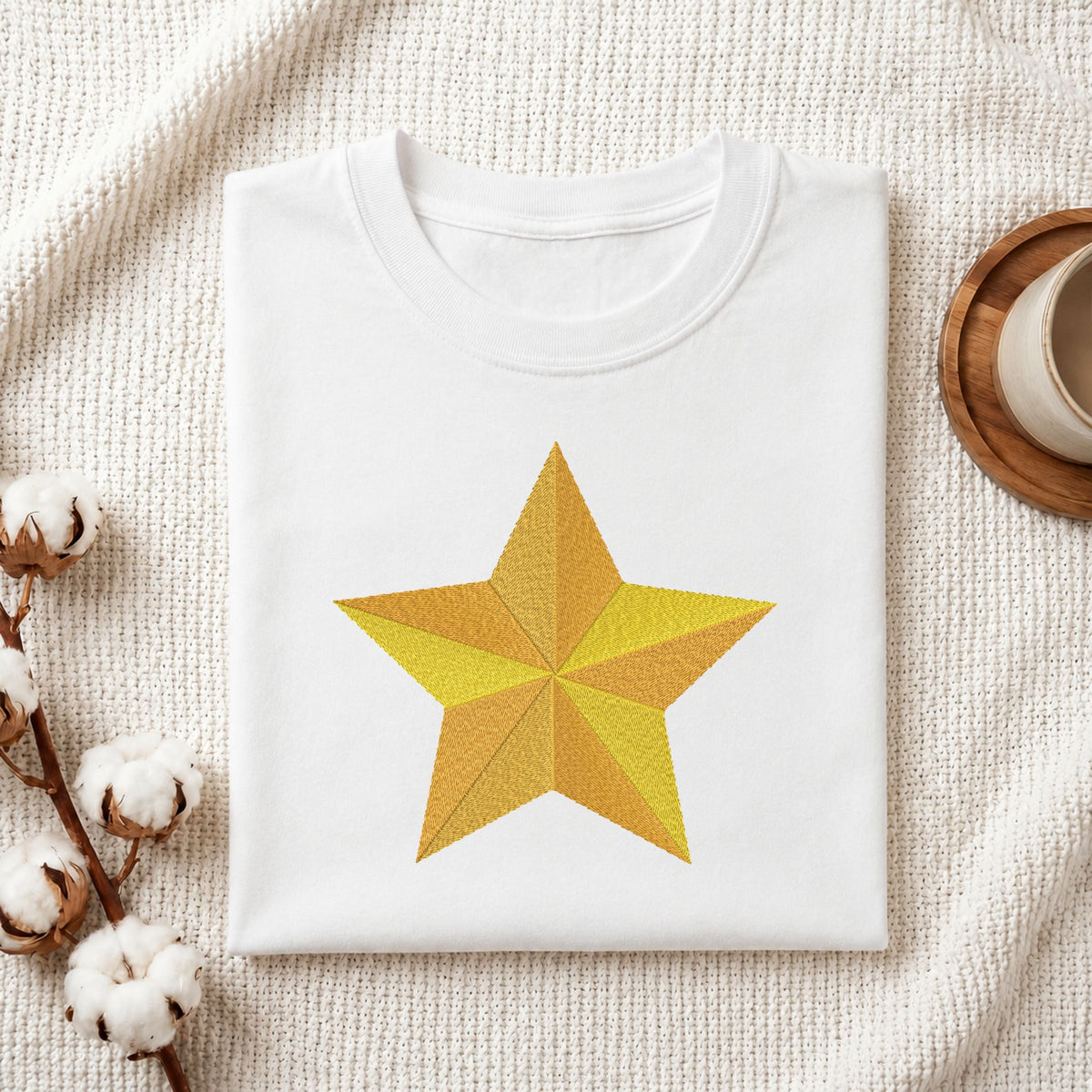 Folded white cotton t-shirt mockup featuring a segmented golden star machine embroidery design, styled with cotton stems and a wooden cup