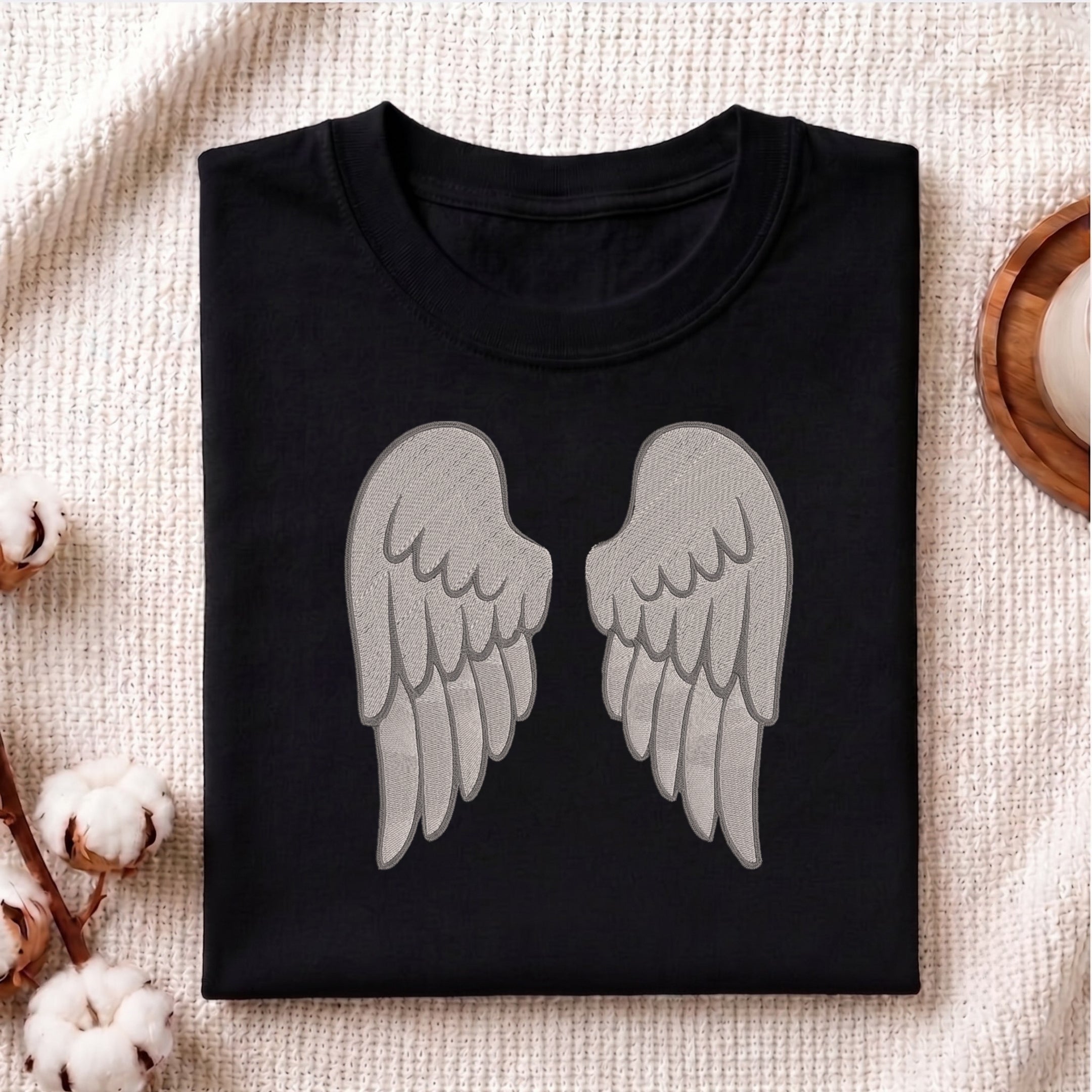 A folded black crewneck t-shirt featuring a large gray embroidered pair of angel wings on the front, styled flat on a textured beige fabric with cotton bolls and a wooden coaster.