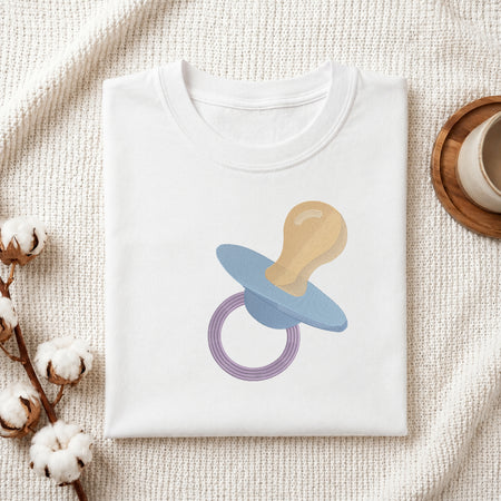 A folded white baby t-shirt with a stylized pacifier graphic in blue, beige, and purple, displayed on a textured cream knit blanket with cotton bolls and a cup in the background.