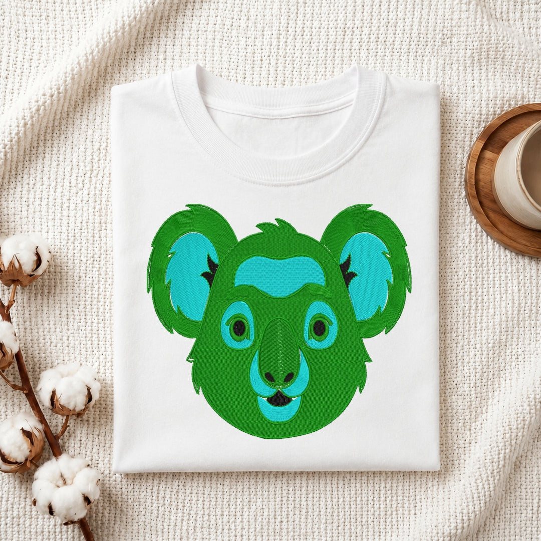 A folded white crewneck t-shirt featuring a colorful graphic of a koala head in shades of green and blue, styled on a textured cream fabric with cotton bolls and a wooden cup.