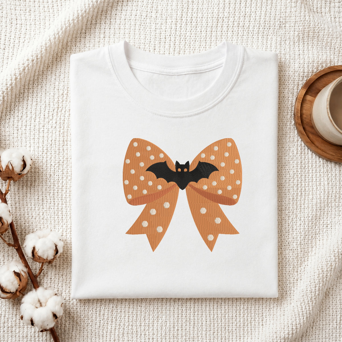 A neatly folded white t-shirt featuring the embroidered orange bat bow design, displayed on a textured white knit background with cotton stems and a wooden cup.