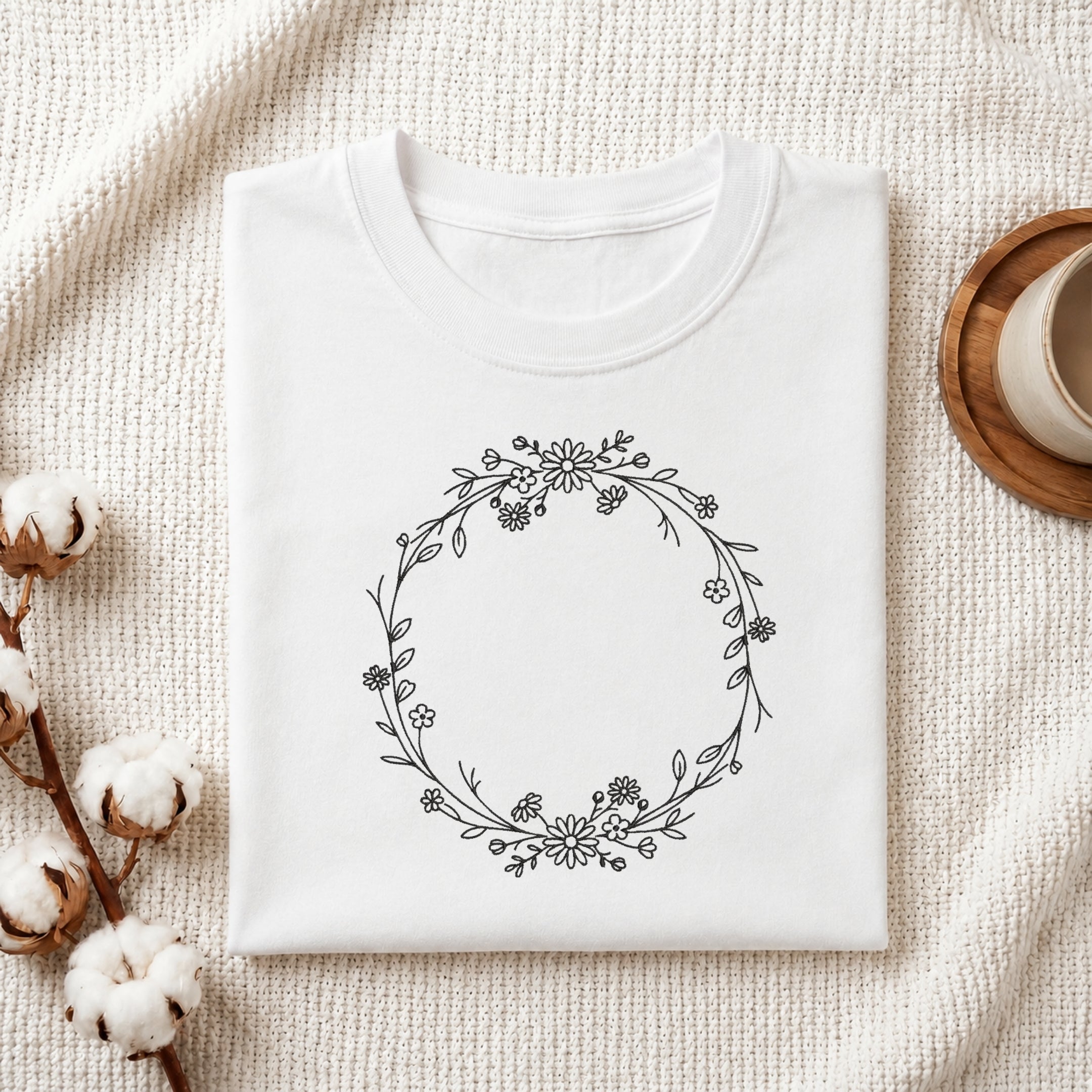 A folded white crewneck t-shirt with a black floral wreath graphic on the front, styled on a textured cream fabric with cotton bolls and a wooden cup in the background.