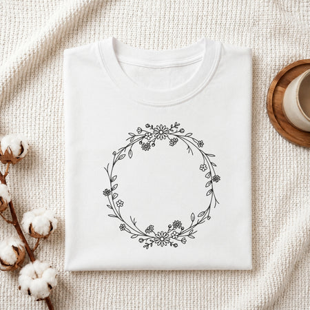A folded white crewneck t-shirt with a black floral wreath graphic on the front, styled on a textured cream fabric with cotton bolls and a wooden cup in the background.