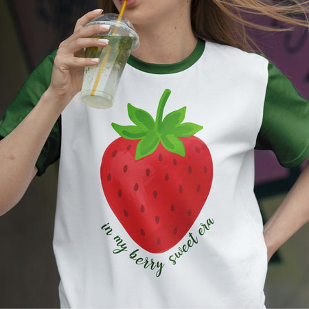 Person wearing a white t-shirt with a strawberry graphic and text, holding a drink.