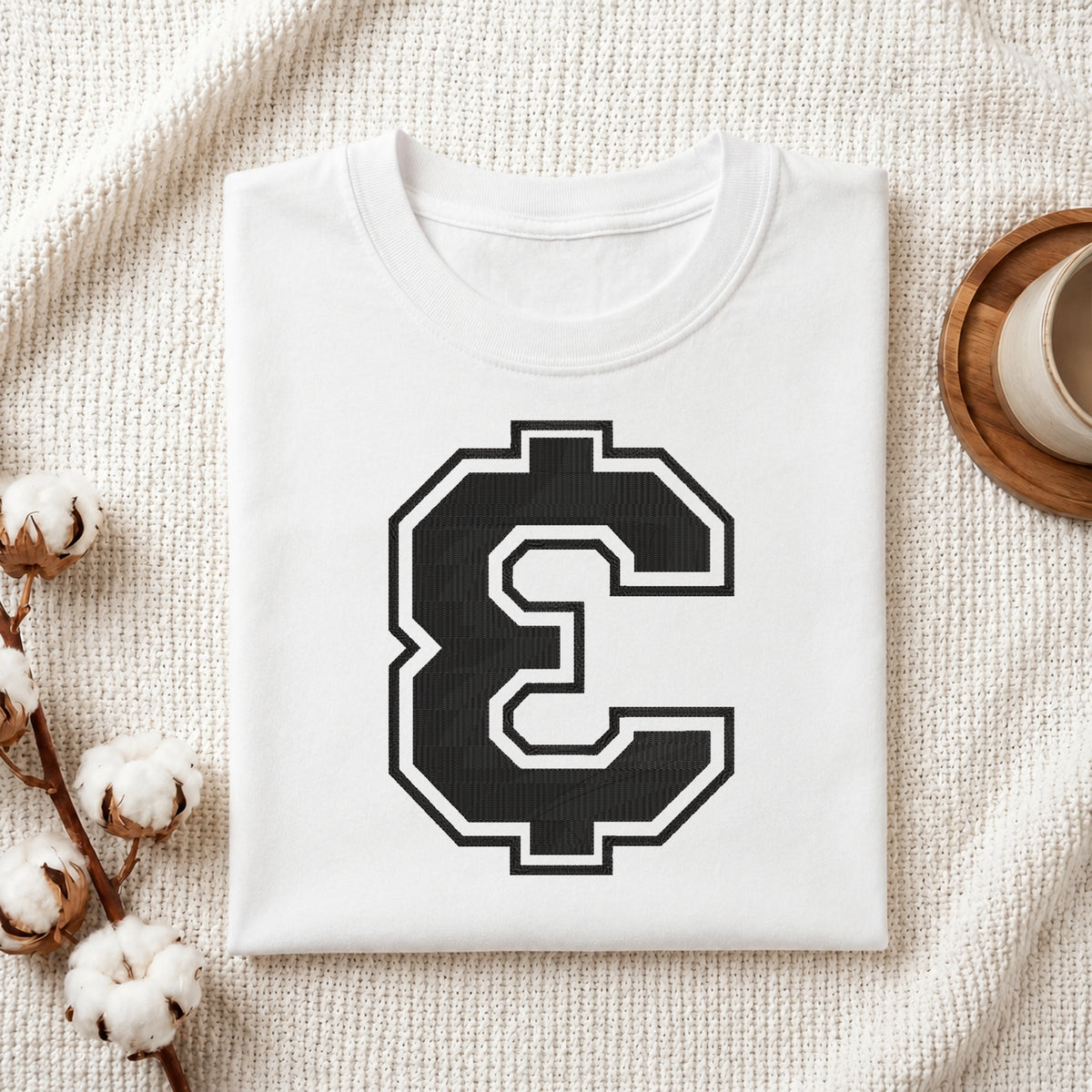 A folded white crewneck t-shirt with a large black and white block letter 'C' graphic on the front, styled on a textured cream knit blanket with cotton bolls and a wooden cup in the background.