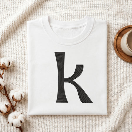 A folded white t-shirt with a large black textured letter 'k' printed on the front, styled on a beige woven blanket with cotton stems and a wooden cup in the background.