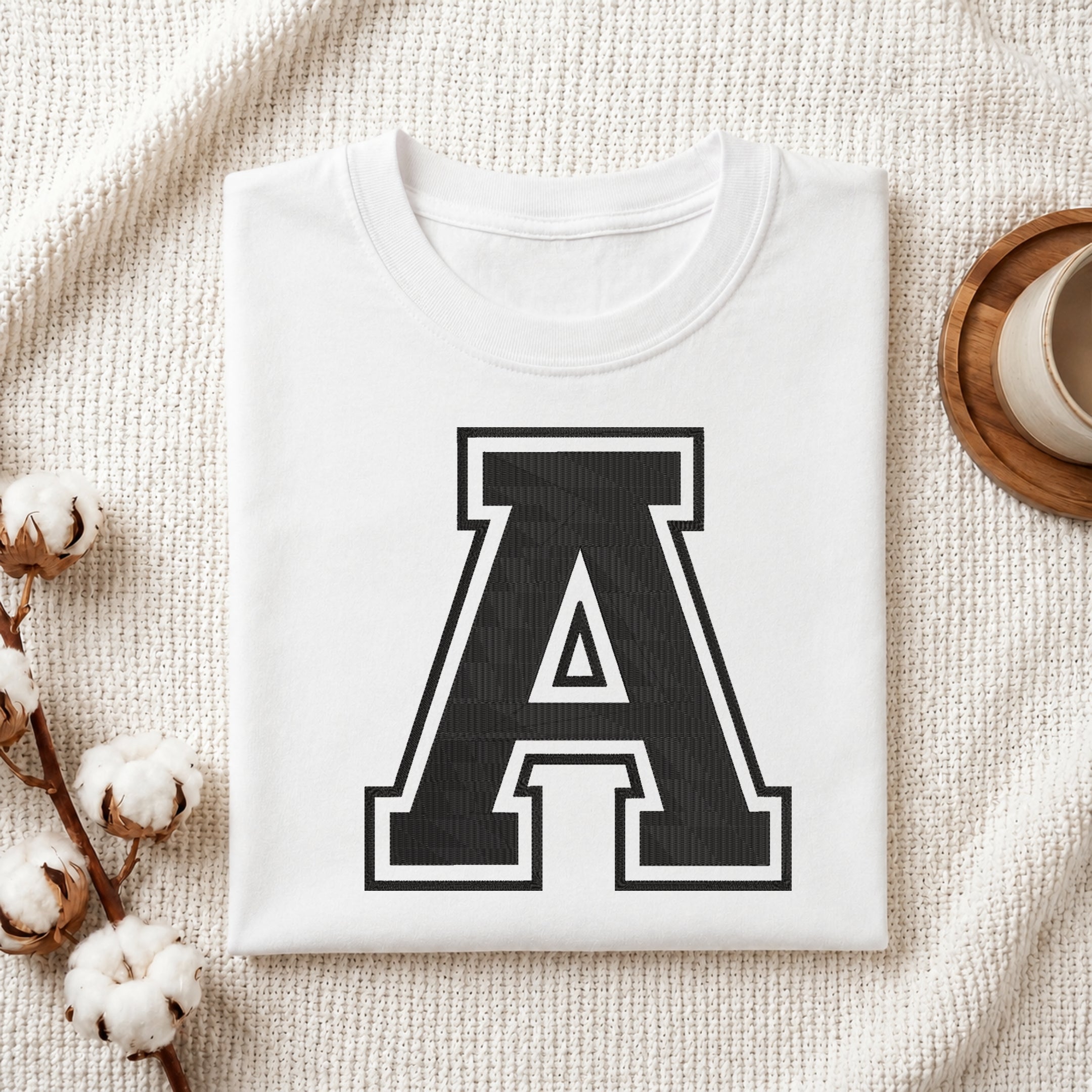 A folded white crewneck t-shirt with a large black collegiate letter 'A' printed on the front, styled in a flat lay on a textured cream fabric with cotton bolls and a cup on a wooden coaster.