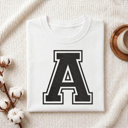 A folded white crewneck t-shirt with a large black collegiate letter 'A' printed on the front, styled in a flat lay on a textured cream fabric with cotton bolls and a cup on a wooden coaster.