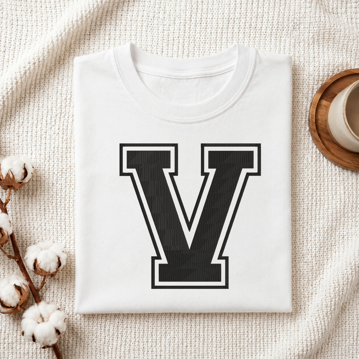 A person carrying a white canvas tote bag featuring a large embroidered varsity block letter 'V' in black with a white outline, set in a bright room with green house plants.